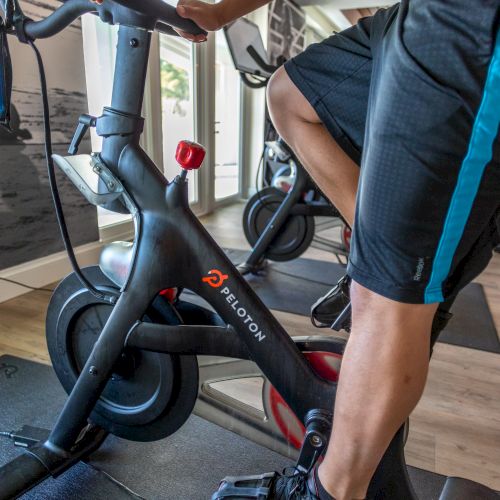 A person is exercising on a Peloton stationary bike indoors, with windows in the background.