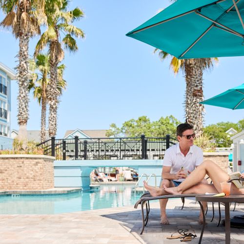 A man and woman relaxing by a pool under umbrellas, surrounded by palm trees and buildings on a sunny day.