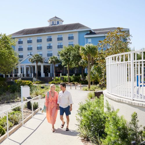 A couple is walking along a landscaped path near a large hotel with several stories and prominent columns.
