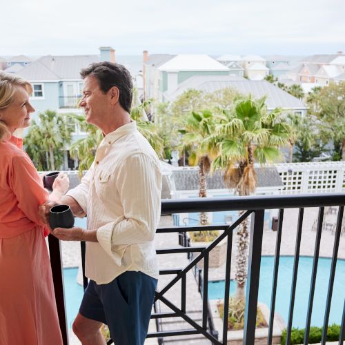 A couple stands on a balcony holding mugs, with a view of a pool, palm trees, and pastel-colored houses in the background.