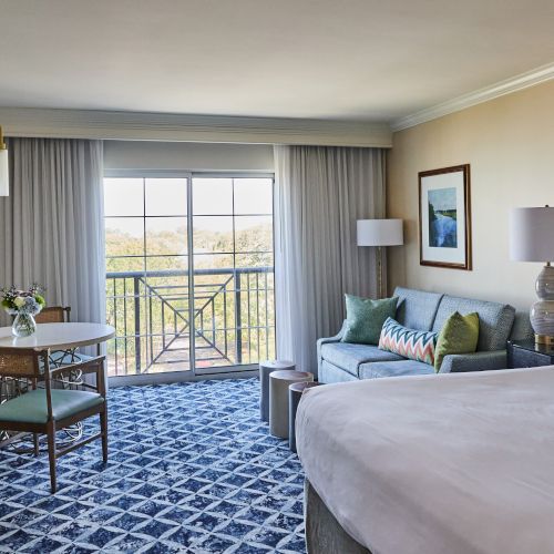 A hotel room with a bed, sofa, table, chairs, lamps, and art, featuring a window with a view of greenery and a balcony.