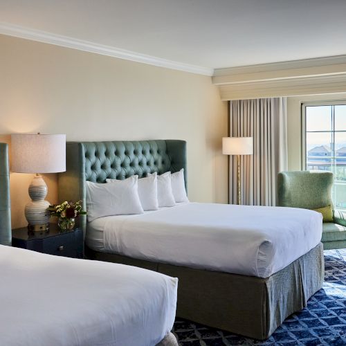 A hotel room with two beds, a lamp, armchair, and balcony view, featuring elegant decor and natural light from the window.