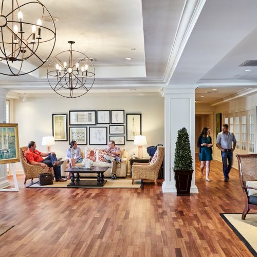 A spacious lobby with people sitting and walking on wooden flooring, adorned with chandeliers, paintings, and potted plants.