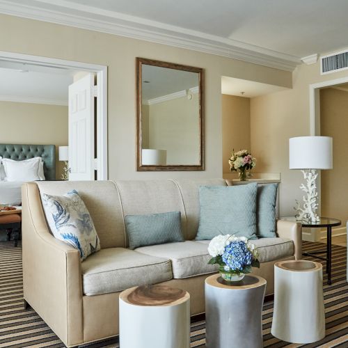A stylish hotel room with a sofa, chairs, pillows, a bed, flowers, and a lamp on a glass table in a cozy setting.