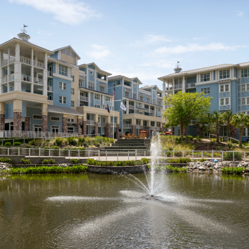 A large residential building complex with a pond and fountain in front, surrounded by greenery and pathways under a clear sky.