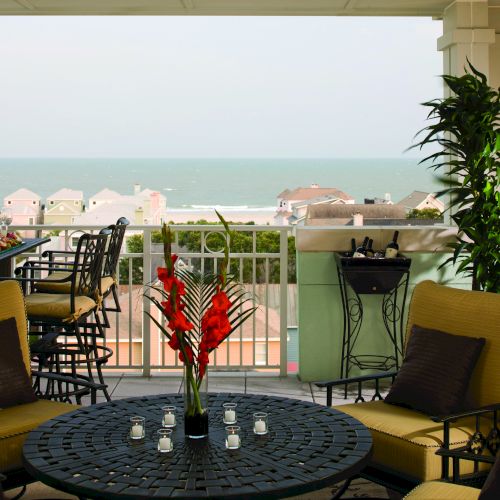 Cozy patio with ocean view, seating, table with flowers and candles, bar with drinks, and tall plant on the right side of the image.