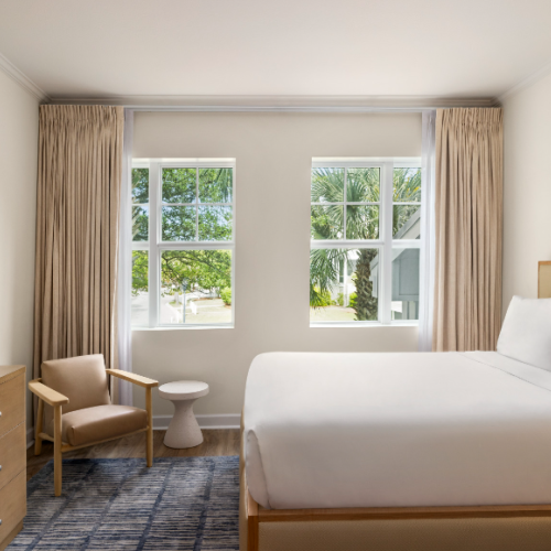 A bright, tidy hotel room with a bed, side chair, TV, dresser, and a window view of trees. Art hangs on the wall above the bed.
