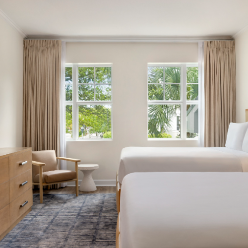 A hotel room with two beds, a TV, dresser, chair, and windows with curtains, overlooking greenery outside.