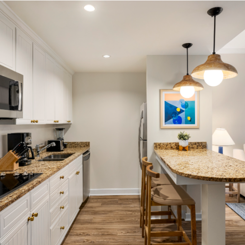 A modern kitchen with white cabinets, granite countertops, bar seating, and hanging lights; adjacent cozy living area with artwork.