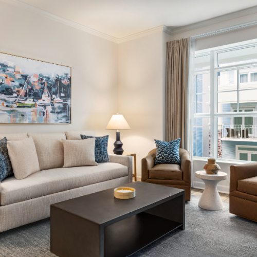 A cozy living room with a beige sofa, two brown armchairs, modern art, and large windows filling the space with natural light.