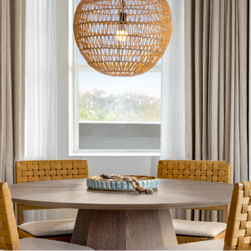 A cozy dining area with a round wooden table, four woven chairs, and a rattan pendant light above. Neutral curtains frame the window scene.