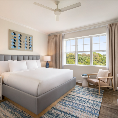 A cozy bedroom with a large bed, nightstand, and lamps. There's a window with curtains, a chair, and a TV. The decor is modern.