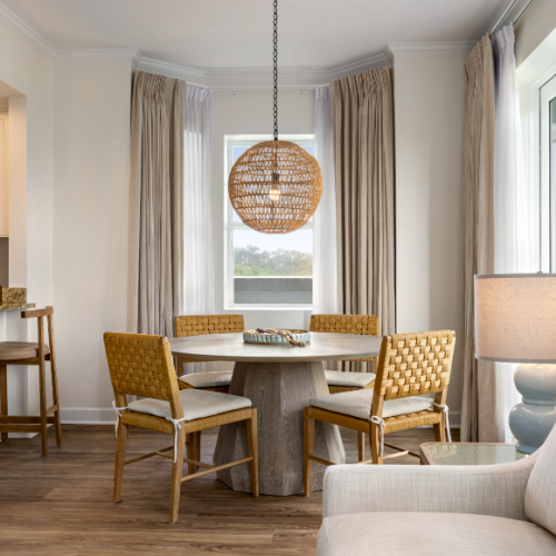 Cozy dining area with wicker chairs and round table, next to a kitchen with bar stools; pendant light overhead and window view.
