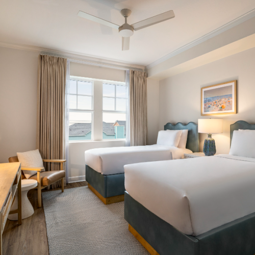 A cozy hotel room with two beds, a desk, a chair, a lamp, a TV, and a window with curtains, under a ceiling fan and lit softly.