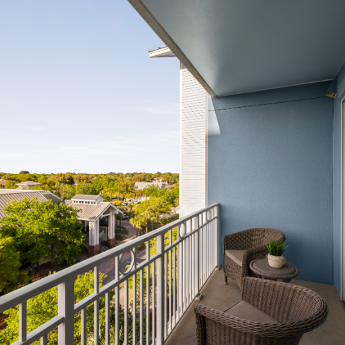 A small balcony with two wicker chairs and a table offers a scenic view of trees and rooftops, surrounded by a blue and white wall.