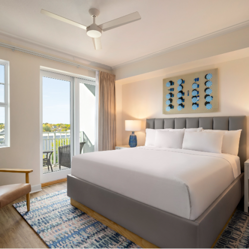 A cozy bedroom with a large bed, two lamps, wall art, a chair, windows, and a balcony view, all in a neutral color palette.