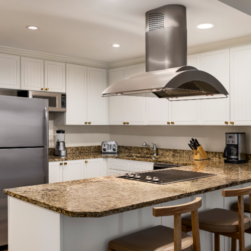 A modern kitchen with granite countertops, stainless steel appliances, white cabinets, and bar stools. An island with a cooker is present.