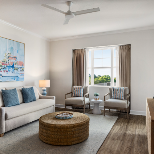 A modern living room with a sofa, chairs, a large window, a ceiling fan, and a wall-mounted TV, featuring neutral tones and tasteful decor.