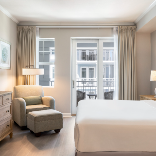 A modern bedroom with a bed, dresser, chair, lamp, and a window view of a building across. Neutral tones create a serene ambiance.