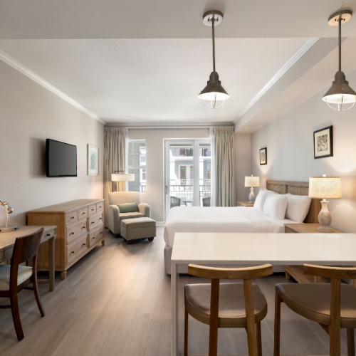 A modern hotel room with two beds, a desk, chair, dresser, TV, and ample lighting, featuring a minimalistic and cozy design.