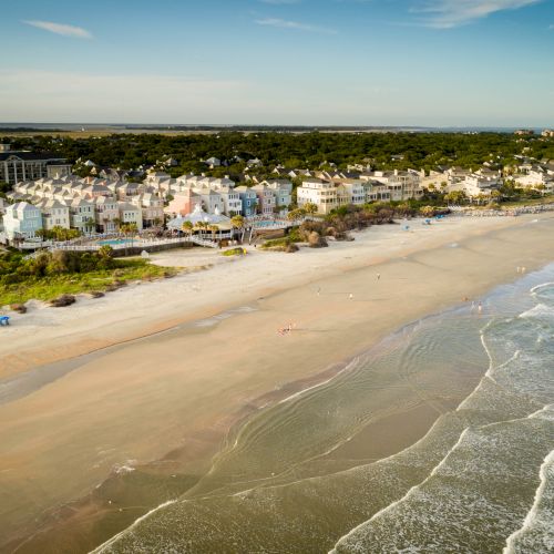 Aerial view of a coastal beach with colorful beachfront houses and people walking along the shore under a clear blue sky.