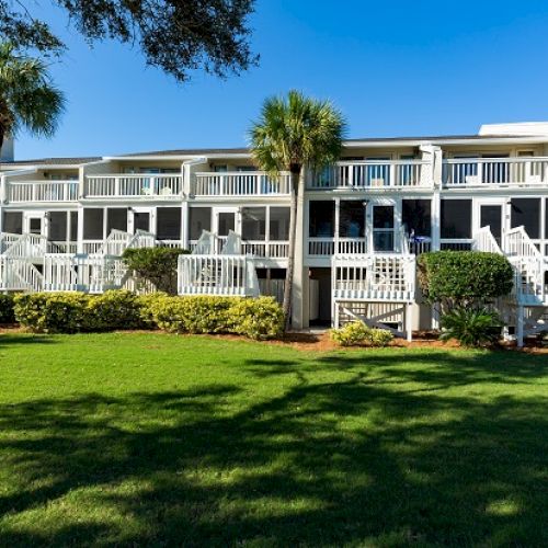The image shows a white building with balconies, surrounded by palm trees and a well-maintained lawn under a clear blue sky.
