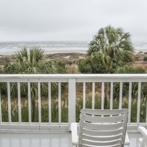 A beachfront view from a balcony featuring a white chair and palm trees, overlooking the ocean under a cloudy sky.