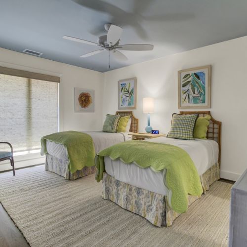 A bedroom with two twin beds, green bedding, a ceiling fan, artwork, wicker chair, and a dresser with a TV remote.