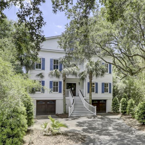A white house with blue shutters, surrounded by lush greenery and palm trees, featuring a double staircase leading to the entrance.