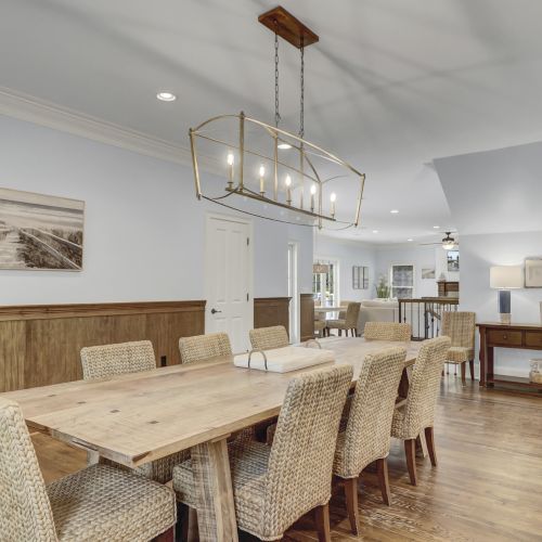 A dining room with a wooden table, wicker chairs, and a modern light fixture, connected to a living area with a staircase.