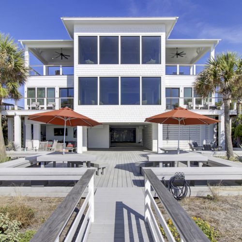 The image shows a modern beach house with large windows, palm trees, red umbrellas, and a wooden deck leading to it.