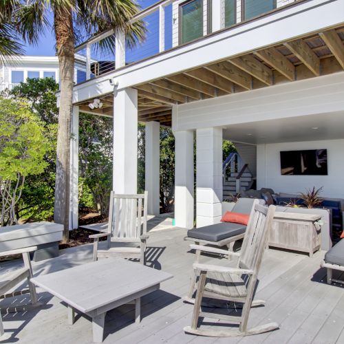 A modern outdoor patio with wooden furniture, rocking chairs, and lounge chairs under a covered area. There's a TV and plants for decor.