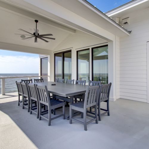 A covered patio with a large dining table, several chairs, ceiling fan, and ocean view is shown.