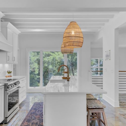 A modern kitchen with white cabinets, a marble island, wicker pendant lights, and large windows overlooking greenery completes the scene.