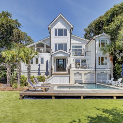 A large, modern white house with multiple floors, surrounded by trees, features a pool and lounge chairs on a grassy yard.