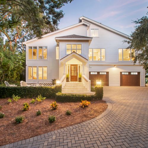 A modern, two-story house with large windows, situated among trees, featuring a landscaped garden and a paved driveway at dusk.