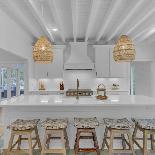 Modern white kitchen with large island, wicker pendant lights, wooden stools, and a view of greenery through the windows.