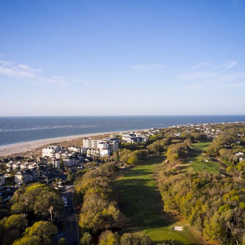 Aerial view of a coastal area with buildings near the beach, a golf course, and surrounding greenery under a clear blue sky.