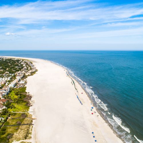 Aerial view of a coastline with sandy beach, ocean waves, and seaside homes. Beach umbrellas line the shore under a clear blue sky.