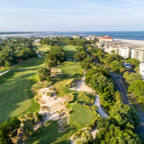 Aerial view of a golf course near the ocean, with trees, a road, and buildings near the coastline.