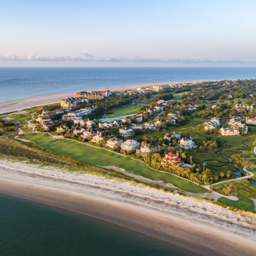 Aerial view of a coastal community with houses, greenery, and a sandy beach meeting the ocean under a clear sky.