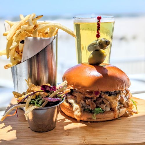 A gourmet burger topped with olives, alongside fries and slaw in cups on a wooden board, with a drink in the background.