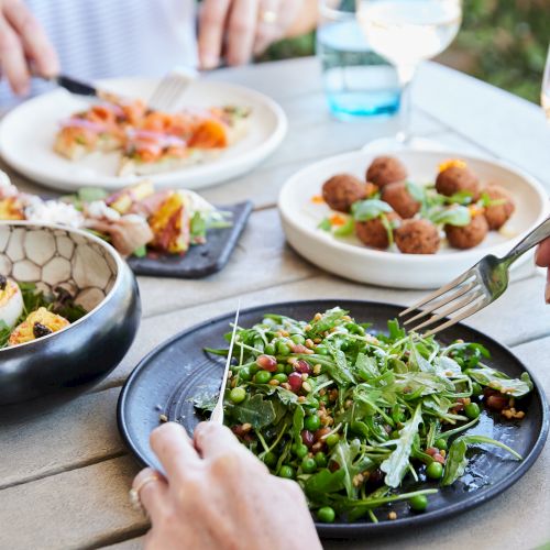 A table with various dishes: a salad with greens and peas, deviled eggs, croquettes, and sliced salmon, accompanied by wine glasses.