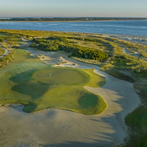 A coastal golf course with lush greens, fairways, and sand traps, alongside residential homes and a scenic ocean view in the background.