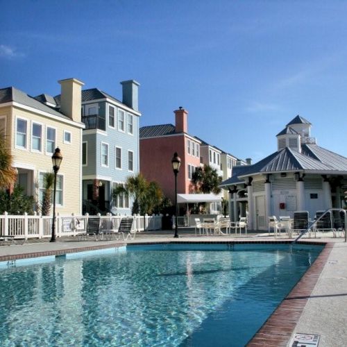 A colorful row of houses lines a sunny outdoor pool area, with lounge chairs and palm trees creating a relaxing atmosphere.