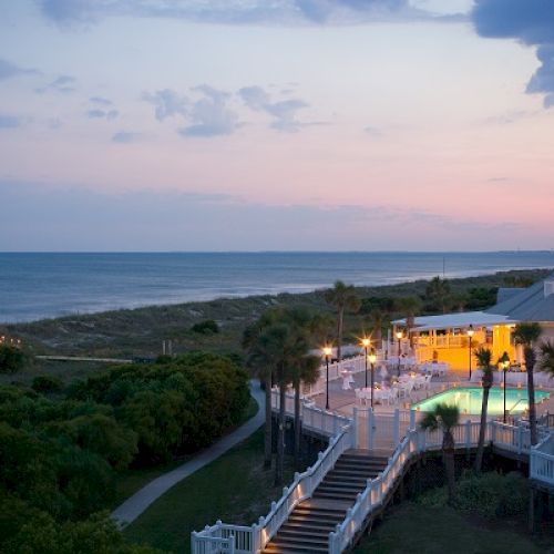 A scenic view of a beachfront resort at dusk, featuring a lit pool area and a pathway leading to the ocean.