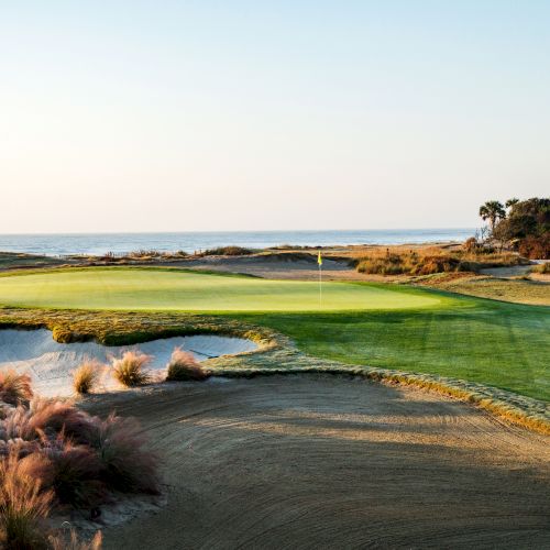 A coastal golf course with green fairways, sand bunkers, ocean view, and buildings in the distance, under a clear sky.