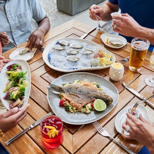 A table with plates of oysters, a grilled fish, salad, cocktails, and beer, surrounded by people enjoying the meal.