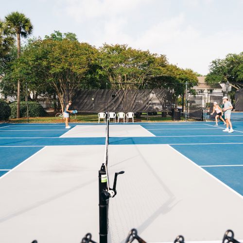 People playing pickleball on a blue court surrounded by trees, with chairs and a bicycle in the background.