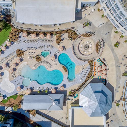 Aerial view of a resort with multiple swimming pools, sun loungers, umbrellas, and surrounding modern buildings.